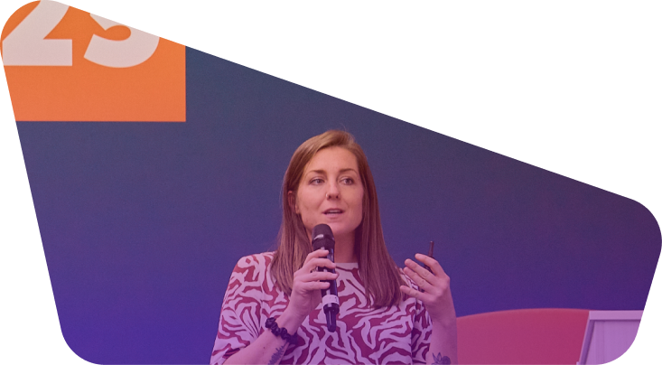 Woman with long brown hair speaking into microphone at conference with purple backdrop